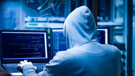 Cybersicherheit: Ein Mensch mit grauem Kapuzenpullover sitzt in einem blau beleuchtetem Raum vor einem Computer und gibt etwas ein.