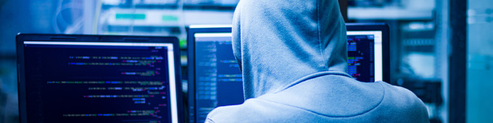 Cybersicherheit: Ein Mensch mit grauem Kapuzenpullover sitzt in einem blau beleuchtetem Raum vor einem Computer und gibt etwas ein.