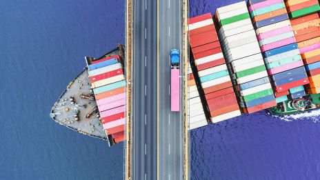 Ein Container-Frachtschiff fährt unter einer großen Brücke hindurch. Von oben ist ein LKW zu sehen, der über die Brücke fährt und die bunten Container auf dem Schiff. 