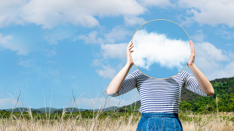 Cloud: Frau steht mit Spiegel vor dem Gesicht in einem Feld. Im Spiegel spiegeln sich die Wolken.