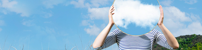 Cloud: Frau steht mit Spiegel vor dem Gesicht in einem Feld. Im Spiegel spiegeln sich die Wolken.