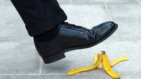 Schwarzer Männerschuh in Schrittbewegung kurz oberhalb einer Bananenschale, die auf dem Boden liegt.