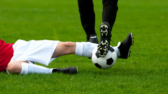 Ein Fußballer in einem roten Trikot rutscht mit gestrecktem Bein in den Ballführenden Fußballer mit schwarzem Trikot rein und spielt dadurch Foul. 