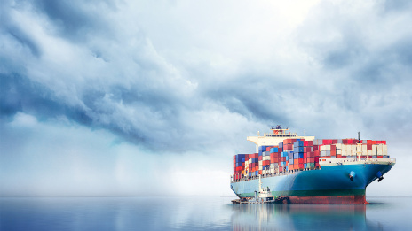 Ein Frachtschiff ist auf Meer zu sehen. Die bunten Container spiegeln sich im Wasser. Vor dem Schiff tut sich eine graue Wolkenwand auf. 
