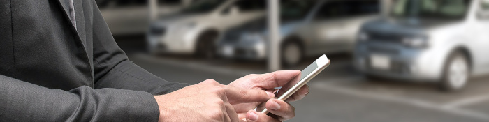 Im Vordergrund ist eine Hand zu sehen, die auf einem Smartphone tippt. Im Hintergrund sind fünf parkende Autos zu sehen. 