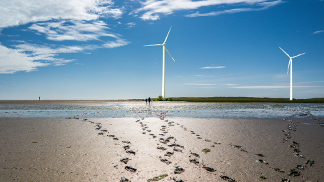 ESG: Windräder stehen im Wattenmeer, im Vordergrund Sandstrand bei Ebbe mit Fußabdrücken im Sand
