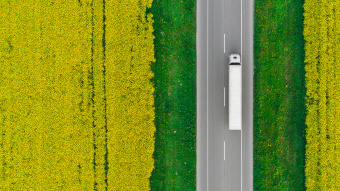 Eine Luftaufnahme zeigt einen LKW auf einer Straße fahrend. Die Straße wird von einzelnen Bäumen gesäumt; ringsherum ist hellgrüne Landschaft zu sehen.