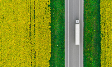 Eine Luftaufnahme zeigt einen LKW auf einer Straße fahrend. Die Straße wird von einzelnen Bäumen gesäumt; ringsherum ist hellgrüne Landschaft zu sehen.