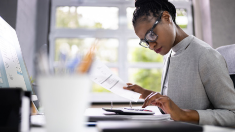 Eine Frau in einem hellgrauen Blazer sitzt an einem Schreibtisch mit Bildschirm und tippt in einen Taschenrechner, in der rechten Hand ein Blatt Papier. 