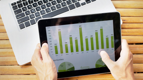 Person mit Tablet auf dem verschiedene Grafiken und Zahlen in grüner Farbe abgebildet sind.