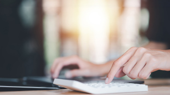 Eine Frauenhand liegt auf der Tastatur von einem Laptop und tippt nebenbei auf einem Taschenrechner.