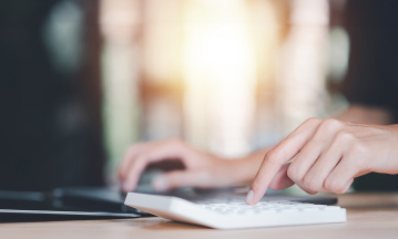 Eine Frauenhand liegt auf der Tastatur von einem Laptop und tippt nebenbei auf einem Taschenrechner.