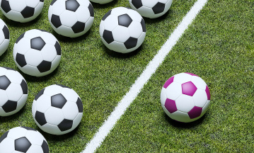 Ein lilafarbener Fußball liegt neben einer Linie, hinter liegen in Reih und Glied schwarz-weiße Fußbälle