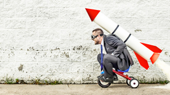 Performance steigern - so kann es klappen. Bildbeschreibung: Erwachsener Mann sitzt auf einem KInderdreirad. Er trägt eine Pilotenbrille. Auf seinen Rücken hat er eine riesige Rakete geschnallt, aus deren Ende Rauch austritt und bald starten wird. 