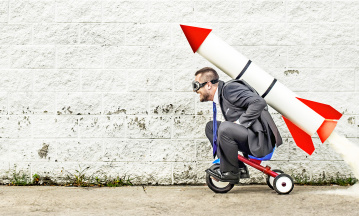 Performance steigern - so kann es klappen. Bildbeschreibung: Erwachsener Mann sitzt auf einem KInderdreirad. Er trägt eine Pilotenbrille. Auf seinen Rücken hat er eine riesige Rakete geschnallt, aus deren Ende Rauch austritt und bald starten wird. 