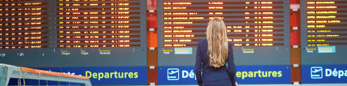 Eine blonde Frau im Business-Outfit steht vor einer Anzeigetafel im Flughafen.