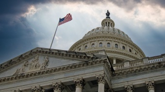 Kamala Harris gegen Donald Trump, US-Präsidentschaftswahl: Das Kapitol in Washington ist zu sehen, im Vordergrund die Flagge der USA, im Hintergrund dunkle Wolken.