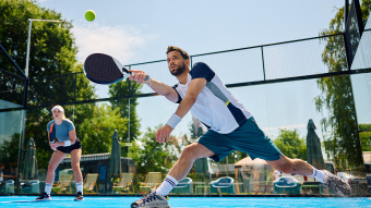 Zwei Padelspieler stehen auf einem Court, der Ball ist im Spiel, im Hintergrund sieht man Grün.