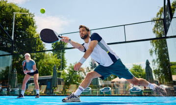 Zwei Padelspieler stehen auf einem Court, der Ball ist im Spiel, im Hintergrund sieht man Grün.