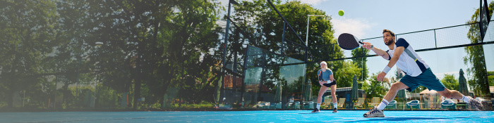 Zwei Padelspieler stehen auf einem Court, der Ball ist im Spiel, im Hintergrund sieht man Grün.