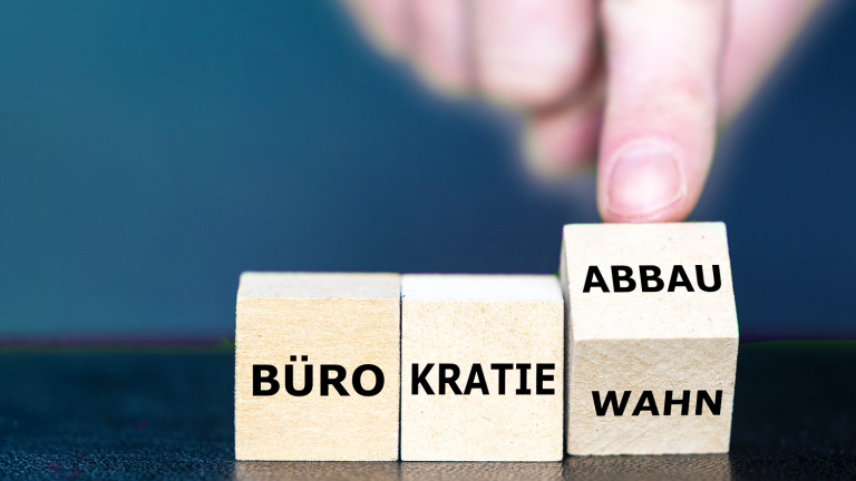 Ein Finger dreht einen Würfel um, auf dem Wahn und Abbau stehen. Auf den beiden links daneben liegenden Würfeln ist das Wort Bürokratie zu lesen.