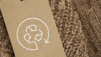Nahaufnahme eines braunen Strickkleidungsstücks mit einem Kartonetikett, das ein weißes Recycling-Symbol zeigt.