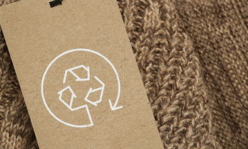 Nahaufnahme eines braunen Strickkleidungsstücks mit einem Kartonetikett, das ein weißes Recycling-Symbol zeigt.