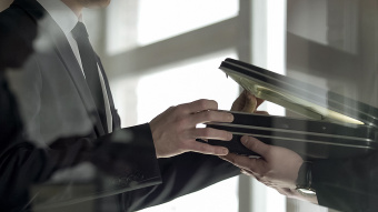 Zwei Männer in Anzügen übergeben sich in einem Büro diskret einen offenen Aktenkoffer – symbolisches Bild für geheime Geschäfte, Korruption oder Wirtschaftskriminalität.