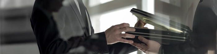Zwei Männer in Anzügen übergeben sich in einem Büro diskret einen offenen Aktenkoffer – symbolisches Bild für geheime Geschäfte, Korruption oder Wirtschaftskriminalität.