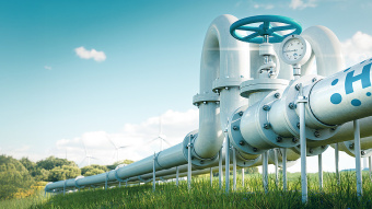 Eine große Wasserstoffpipeline mit einem blauen Ventil und einem Manometer verläuft durch ein grünes Feld. Im Hintergrund stehen Windkraftanlagen unter einem klaren blauen Himmel mit vereinzelten Wolken.