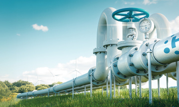 Eine große Wasserstoffpipeline mit einem blauen Ventil und einem Manometer verläuft durch ein grünes Feld. Im Hintergrund stehen Windkraftanlagen unter einem klaren blauen Himmel mit vereinzelten Wolken.