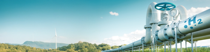 Eine große Wasserstoffpipeline mit einem blauen Ventil und einem Manometer verläuft durch ein grünes Feld. Im Hintergrund stehen Windkraftanlagen unter einem klaren blauen Himmel mit vereinzelten Wolken.