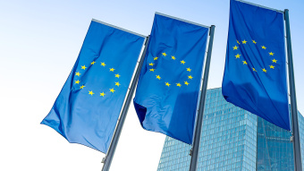 Drei wehende EU-Flaggen vor modernem Glasgebäude bei blauem Himmel – Symbol für die Europäische Union, europäische Politik und internationale Zusammenarbeit in urbanem Umfeld.