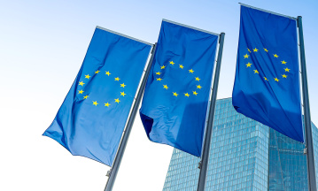 Drei wehende EU-Flaggen vor modernem Glasgebäude bei blauem Himmel – Symbol für die Europäische Union, europäische Politik und internationale Zusammenarbeit in urbanem Umfeld.
