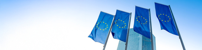 Drei wehende EU-Flaggen vor modernem Glasgebäude bei blauem Himmel – Symbol für die Europäische Union, europäische Politik und internationale Zusammenarbeit in urbanem Umfeld.