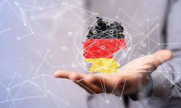 Eine Person im grauen Anzug hält eine offene Hand ausgestreckt. Über der Hand schwebt eine digitale Darstellung der Deutschlandkarte in den Farben der deutschen Flagge (schwarz, rot, gold). Die Karte ist von einem Netzwerk aus Linien und Knotenpunkten umgeben. Der Hintergrund ist unscharf in Blau- und Weißtönen gehalten.