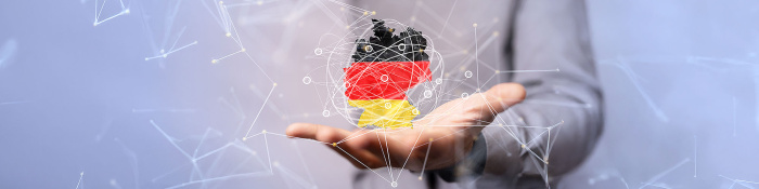 Eine Person im grauen Anzug hält eine offene Hand ausgestreckt. Über der Hand schwebt eine digitale Darstellung der Deutschlandkarte in den Farben der deutschen Flagge (schwarz, rot, gold). Die Karte ist von einem Netzwerk aus Linien und Knotenpunkten umgeben. Der Hintergrund ist unscharf in Blau- und Weißtönen gehalten.