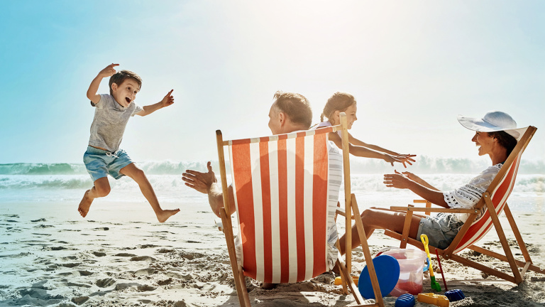 Strandbild mit rot-weiß gestreiftem Sonnenstuhl, Familie mit zwei Kindern, kleiner Junge springt, Eltern sitzen auf Strandstühlen
