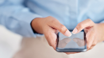 Eine Person hält ein Smartphone in beiden Händen und tippt darauf. Die Person trägt ein hellblaues Hemd.