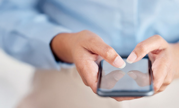 Eine Person hält ein Smartphone in beiden Händen und tippt darauf. Die Person trägt ein hellblaues Hemd.