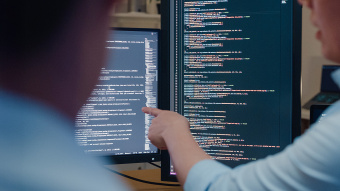 Zwei Computermonitore mit Schrift im Hintergrund, eine Person, von der nur ein Arm zu sehen ist, zeigt mit dem Finger auf einen Monitor, außerdem im Vordergrund noch eine weitere unscharfe Person von hinten (Ausschnitt) 