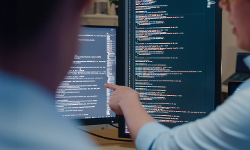 Zwei Computermonitore mit Schrift im Hintergrund, eine Person, von der nur ein Arm zu sehen ist, zeigt mit dem Finger auf einen Monitor, außerdem im Vordergrund noch eine weitere unscharfe Person von hinten (Ausschnitt) 