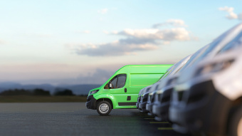 Ein leuchtend grüner Lieferwagen steht in einer Reihe weißer Transporter auf einem Parkplatz, im Hintergrund unscharfe Landschaft mit Hügeln und Himmel.