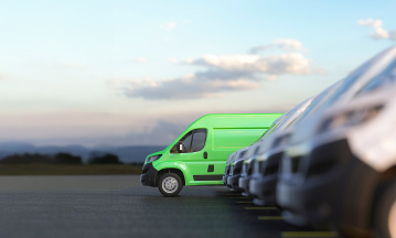 Ein leuchtend grüner Lieferwagen steht in einer Reihe weißer Transporter auf einem Parkplatz, im Hintergrund unscharfe Landschaft mit Hügeln und Himmel.