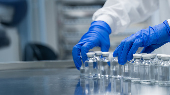 Laborfachkraft mit blauen Schutzhandschuhen richtet mehrere Glasfläschchen mit Flüssigkeit auf einem Edelstahltisch für die Auslaufsteuerung in der pharmazeutischen Produktion aus.