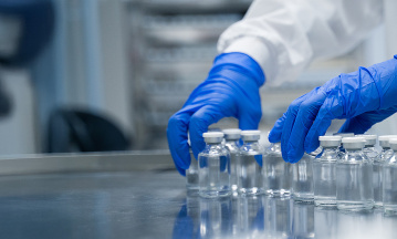 Laborfachkraft mit blauen Schutzhandschuhen richtet mehrere Glasfläschchen mit Flüssigkeit auf einem Edelstahltisch für die Auslaufsteuerung in der pharmazeutischen Produktion aus.
