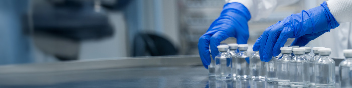 Laborfachkraft mit blauen Schutzhandschuhen richtet mehrere Glasfläschchen mit Flüssigkeit auf einem Edelstahltisch für die Auslaufsteuerung in der pharmazeutischen Produktion aus.