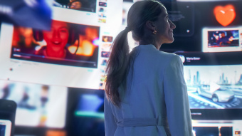 Eine Frau mit blondem Pferdeschwanz steht seitlich vor großen digitalen Bildschirmen, auf denen verschiedene Videos und Symbole zu sehen sind. Unter anderem zu sehen ist ein rotes Herz und ein Porträt einer Person. Die Szene ist in Blautönen gehalten und vermittelt den Eindruck digitaler Vernetzung und Medienvielfalt.