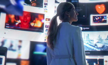 Eine Frau mit blondem Pferdeschwanz steht seitlich vor großen digitalen Bildschirmen, auf denen verschiedene Videos und Symbole zu sehen sind. Unter anderem zu sehen ist ein rotes Herz und ein Porträt einer Person. Die Szene ist in Blautönen gehalten und vermittelt den Eindruck digitaler Vernetzung und Medienvielfalt.