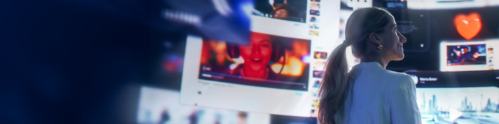 Eine Frau mit blondem Pferdeschwanz steht seitlich vor großen digitalen Bildschirmen, auf denen verschiedene Videos und Symbole zu sehen sind. Unter anderem zu sehen ist ein rotes Herz und ein Porträt einer Person. Die Szene ist in Blautönen gehalten und vermittelt den Eindruck digitaler Vernetzung und Medienvielfalt.